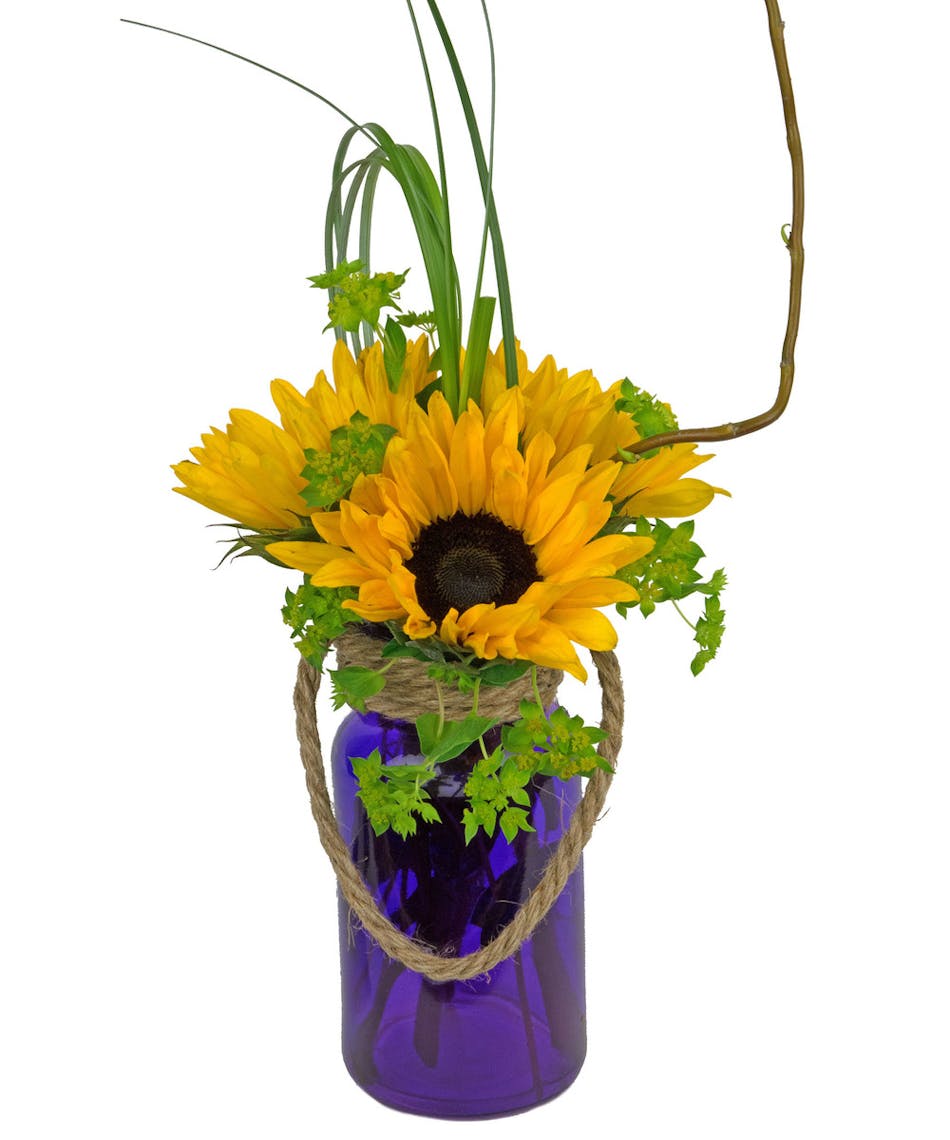 Pretty purple vase is filled with bright sunflowers and other natural looking greenery.