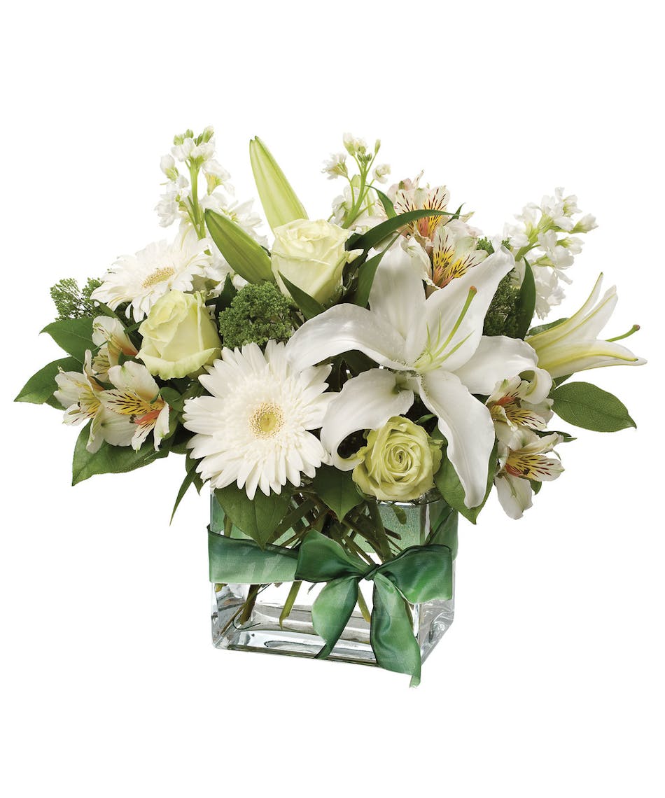 Single white oriental lily surrounded by roses, gerbera daisies, alstromeria and stock in hues of white and green in a clear glass cube vase.