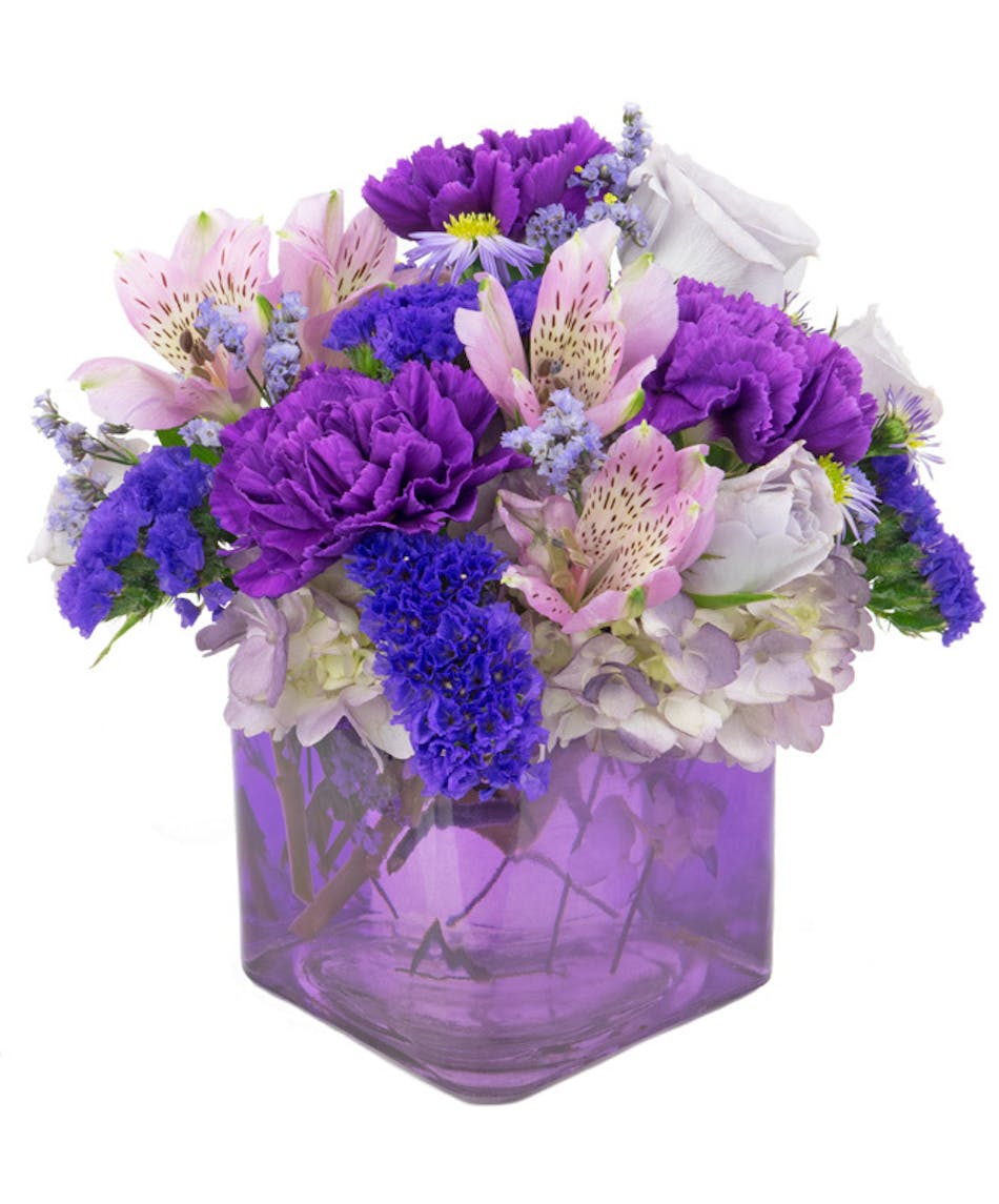 Assorted purple and lavender flowers are arranged in a purple glass cube. 